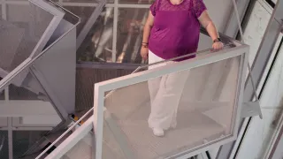 Eine Frau in einem lila Oberteil und weißen Hosen steht auf einer mit Netz überzogenen Treppe und hält sich an das Geländer, mit einem verschwommenen Hintergrund eines Bürogebäude-Inneren.