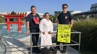 Drei Personen stehen in der Nähe eines Pools, wobei eine Frau in einem weißen Bademantel ein Schild hält und ein Mann ein rotes Herz hält. Ein anderer Mann hält eine schwarze Tasche.
