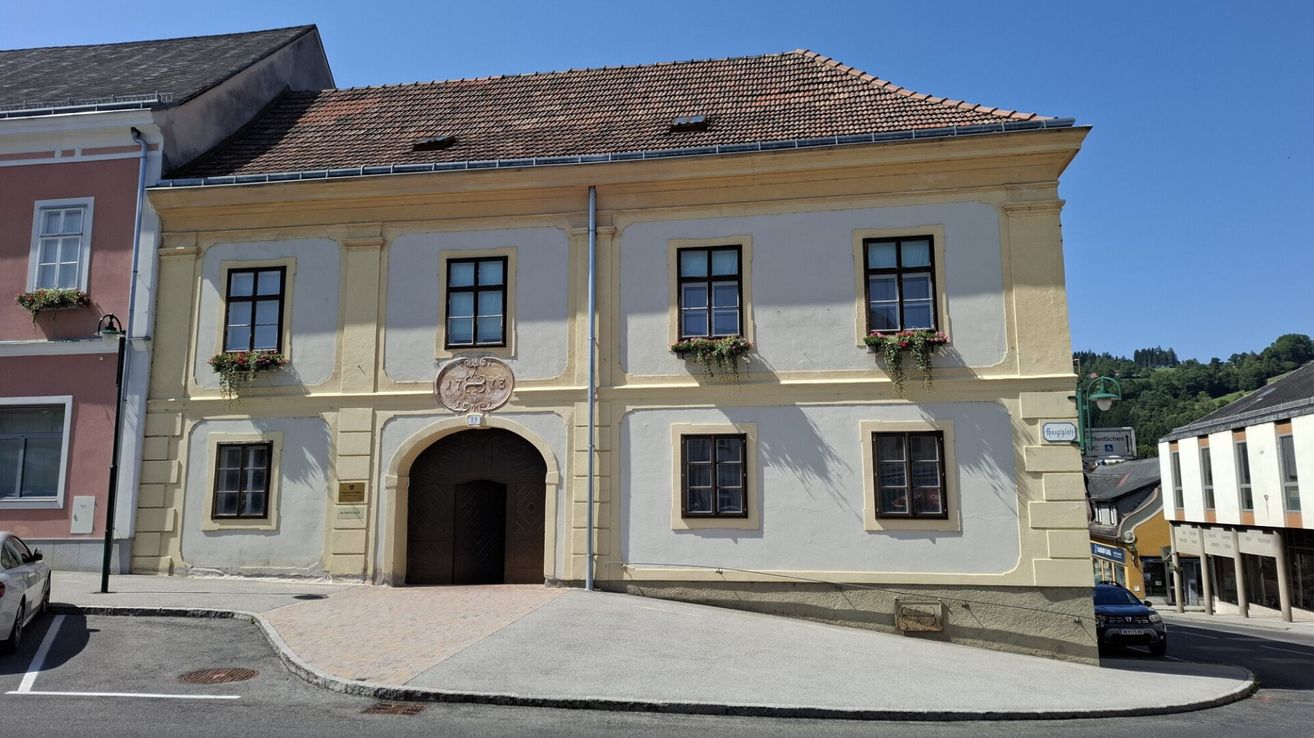 Ein gelbes Gebäude mit einem braunen Bogeneingang hat eine Nummerntafel über ihm und Blumenkästen unter den Fenstern. Das Gebäude hat ein Ziegeldach.