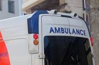 A white ambulance vehicle is parked outside a building with multiple windows. The word 'AMBULANCE' is written on the back of the vehicle.