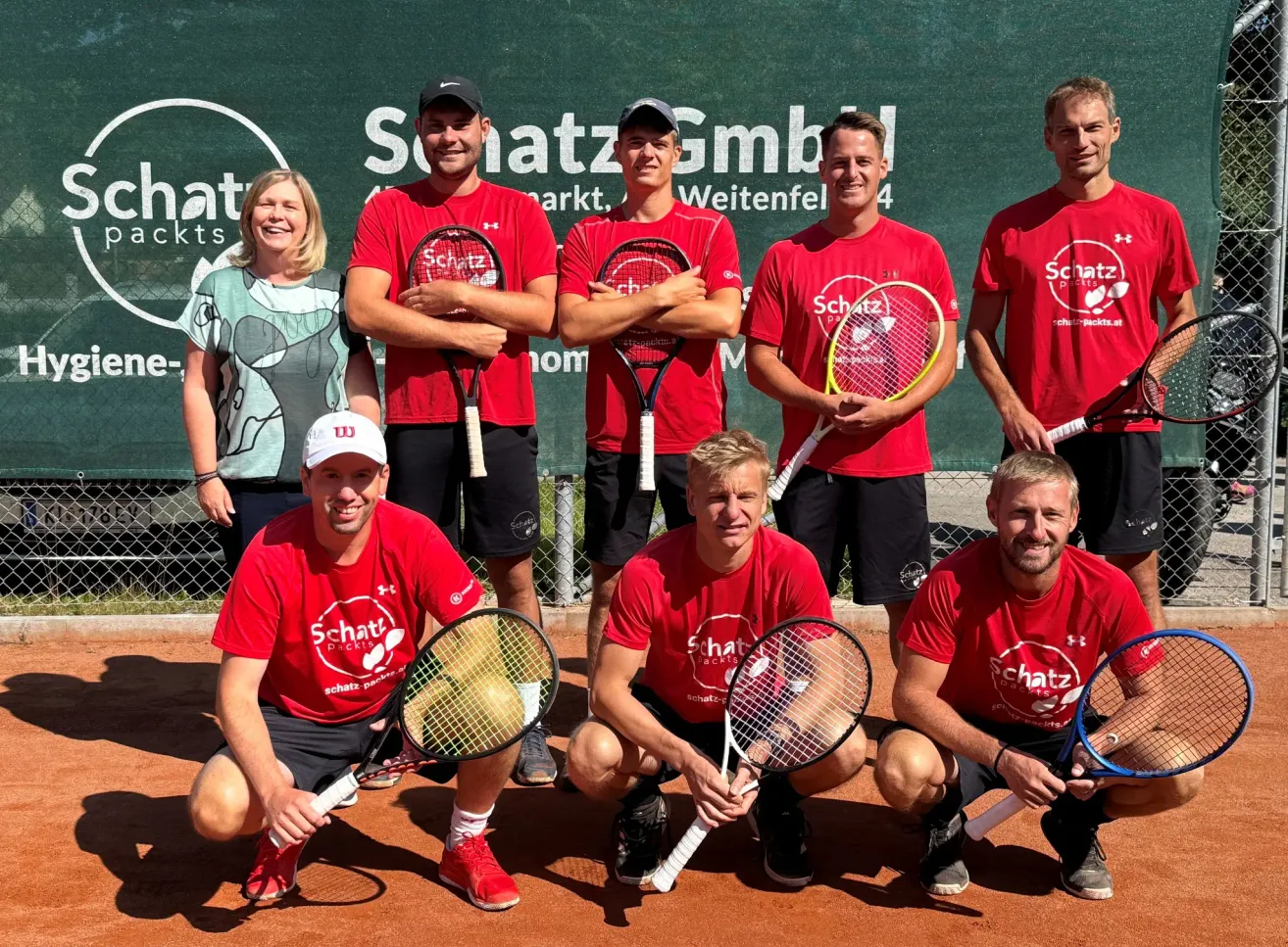 Acht Personen, darunter sechs Männer und zwei Frauen, posieren für ein Foto auf einem Tennisplatz. Alle tragen rote Shirts und halten Tennisschläger. Hinter ihnen befindet sich ein grüner Banner mit weißem Text.