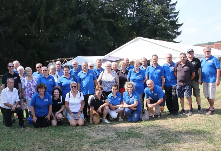 Eine Gruppe von Menschen in blauen Poloshirts, die für ein Foto im Freien posieren, mit Bäumen und einem Zelt im Hintergrund.