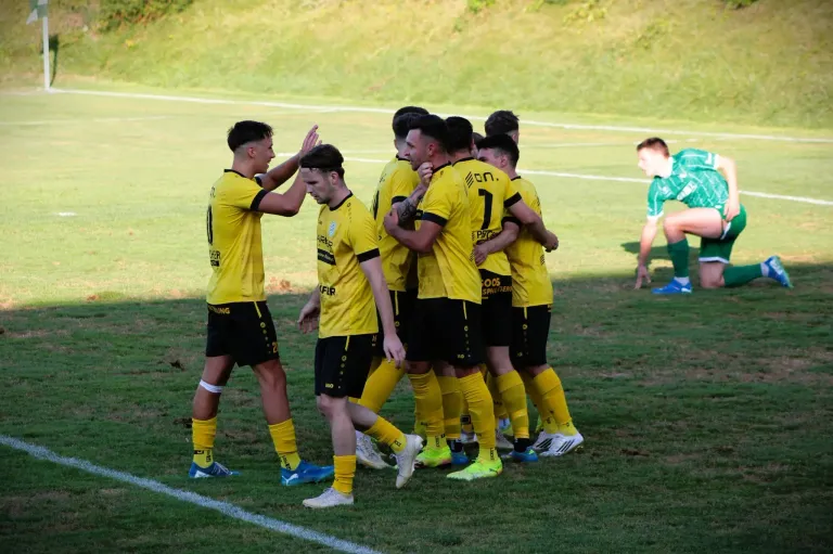 Eine Fußballmannschaft in gelben Trikots feiert ein Tor auf einem Rasenfeld. Sie stehen eng beieinander, ein Spieler in einem grünen Trikot beobachtet von hinten.