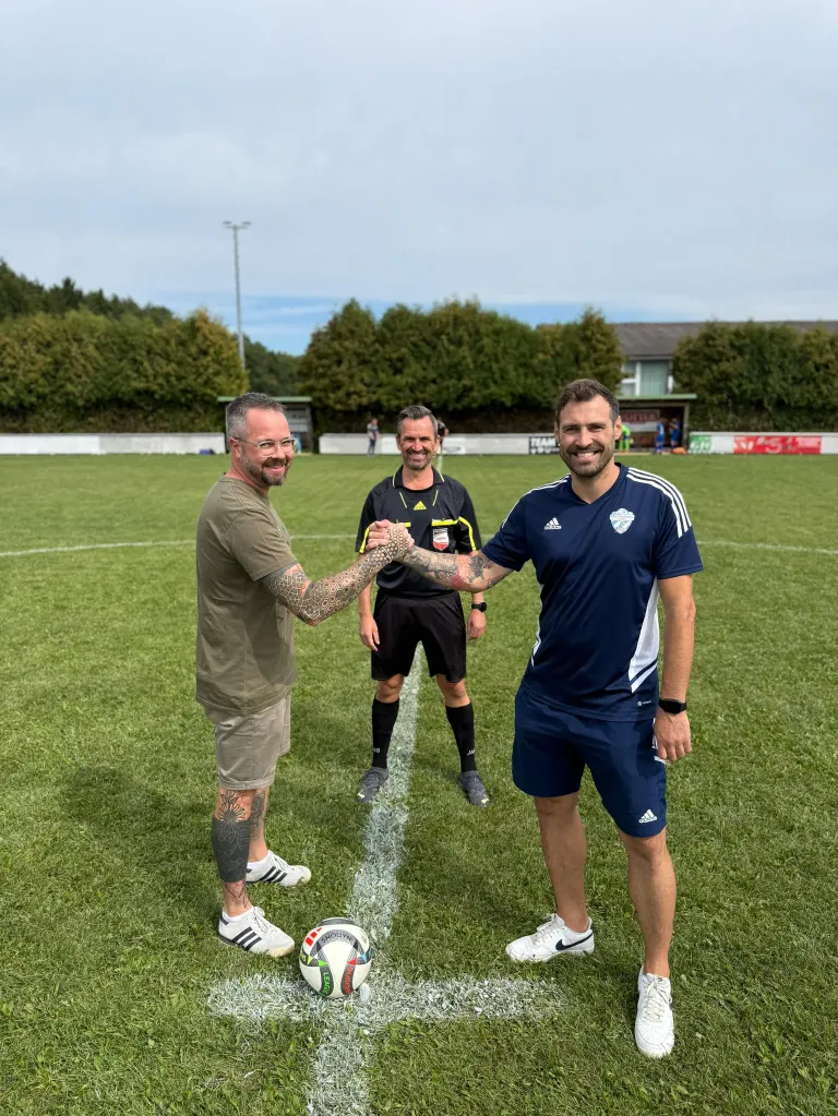 Drei Männer auf einem Fußballfeld. Einer schüttelt dem anderen die Hand, während der dritte in einem schwarz-gelben Trikot hinter ihnen steht. Der Boden ist mit Gras bedeckt, und ein Fußball liegt vor ihnen.