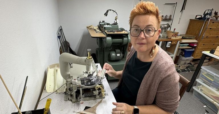 Eine Frau mit rotem Haar sitzt in einer Werkstatt an einer Nähmaschine. Sie trägt eine Brille und einen Ring. Hinter ihr ist eine weitere Maschine zu sehen.