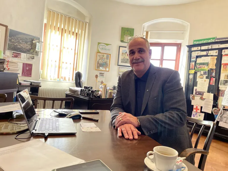 Ein Mann sitzt an einem Schreibtisch mit Laptop und einer Tasse Kaffee, in einem Anzug. Hinter ihm ist ein Fenster und verschiedene Gegenstände zu sehen.