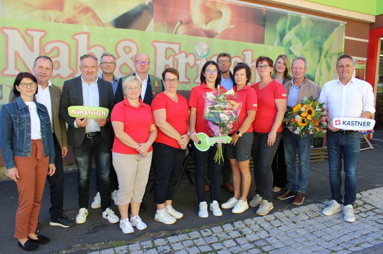 Eine Gruppe von Menschen steht vor einem Banner. Sie tragen alle rote Hemden und halten Blumen. Ein Mann hält ein Schild mit der Aufschrift Nah&Frisch.