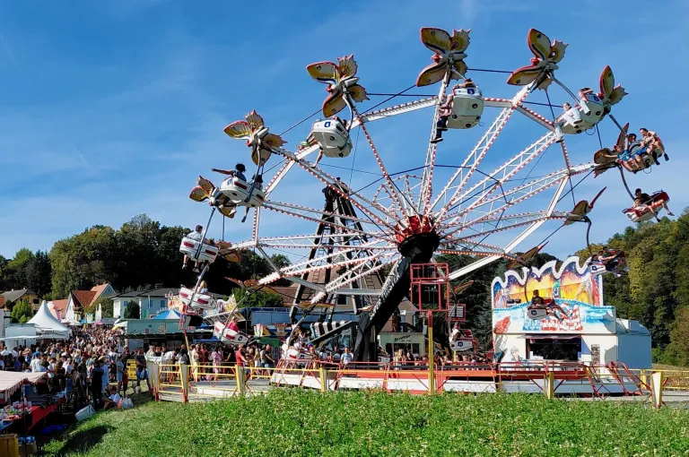A large, colorful Ferris wheel is spinning on a sunny day, with people riding on it and a crowd gathered around.