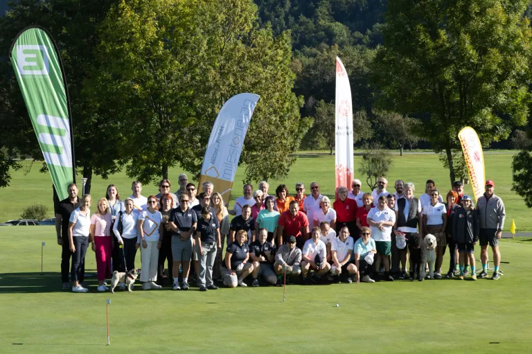 Eine Gruppe von Menschen posiert für ein Foto auf einem Golfplatz mit Bäumen und Bannern im Hintergrund. Einige tragen Hüte und Sonnenbrillen. Es sind Hunde anwesend.