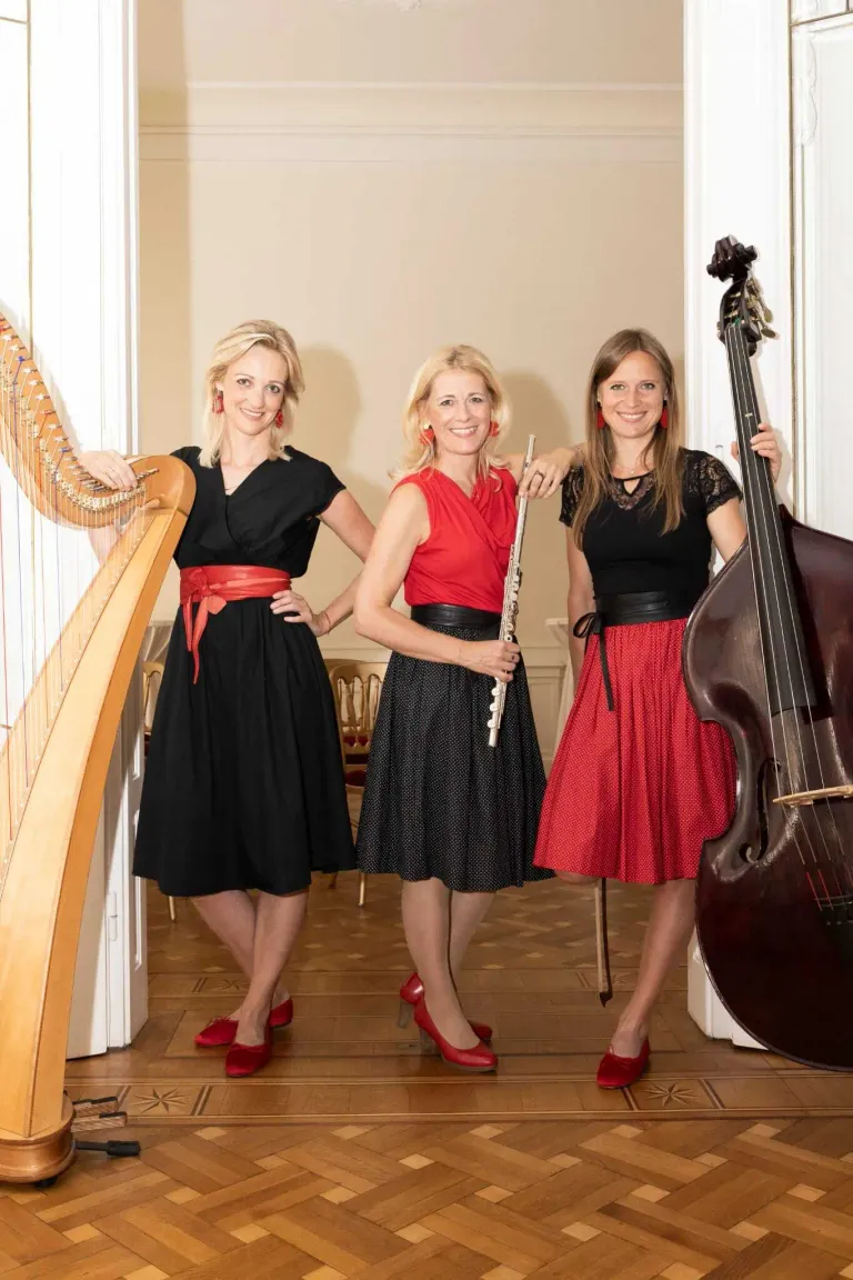 Drei Frauen stehen nebeneinander vor einer weißen Wand. Sie tragen alle rote Ohrringe und rote High Heels. Sie halten alle Musikinstrumente. Die Frau auf der linken Seite hält eine Harfe, die Frau in der Mitte hält eine Flöte und die Frau auf der rechten Seite hält einen Cello.