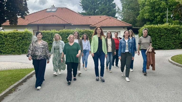 A group of women walks in front of a house with a tiled roof, surrounded by hedges and trees, on a cloudy day.