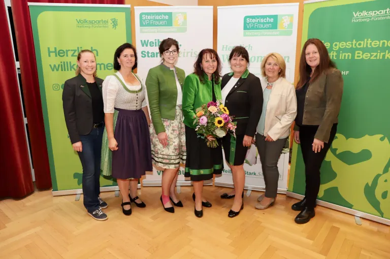 Eine Gruppe von Frauen in formeller Kleidung, darunter traditionelle Kleider, steht vor Bannern mit den Aufschriften 'Steirische VP Frauen' und 'Hartberg-Fürstenfeld', lächelt und posiert für ein Foto.