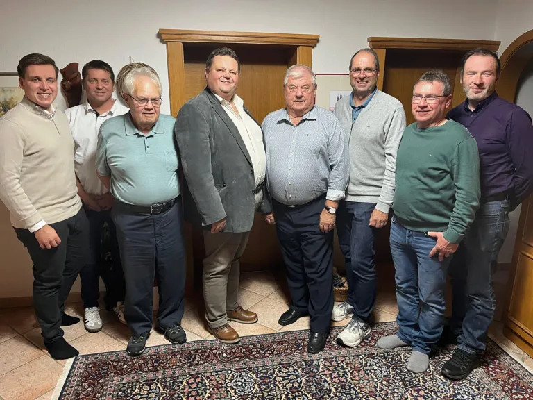 Eine Gruppe von Männern in verschiedenen Kleidungsstücken, die lächelnd für ein Foto in einem Raum mit Holzschränken und einem gemusterten Teppich posieren.