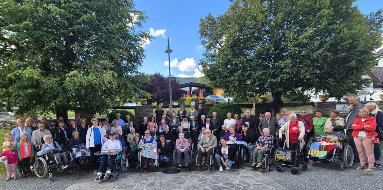 A large group of elderly people, many in wheelchairs, pose for a picture outside in a park. There are trees, plants, and mountains in the background.
