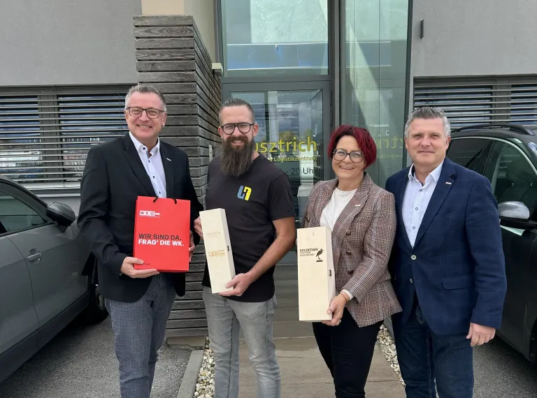 Four people stand outside a building. Three men and one woman. They are holding gift boxes and smiling. The man on the left has a red bag with text. The middle man has a beard and glasses. The woman has short red hair. Behind them is a building with glass doors.