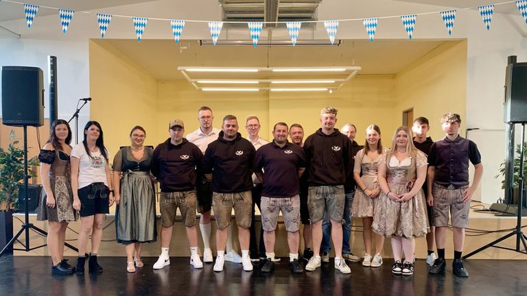 Eine Gruppe von Menschen, gekleidet in traditioneller deutscher Tracht, posiert für ein Foto in einem Raum mit gelben Wänden und blau-weißen Wimpelketten.