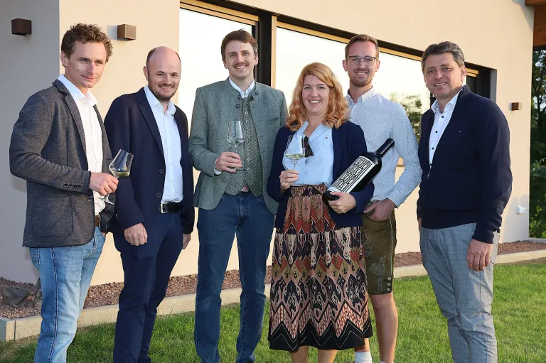 Fünf Personen stehen auf einem Rasen und halten Weingläser und eine Flasche. Sie scheinen für ein Foto zu posieren. Hinter ihnen steht ein Gebäude mit Glasfenstern.