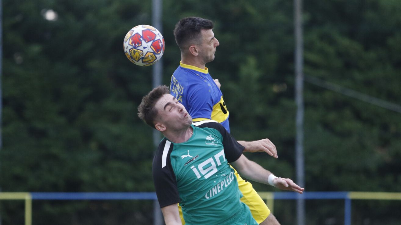 Zwei Fußballspieler in einem Spiel, einer trägt ein blaues Trikot mit gelben Akzenten, der andere in Grün, kämpfen um den Ball in der Luft.