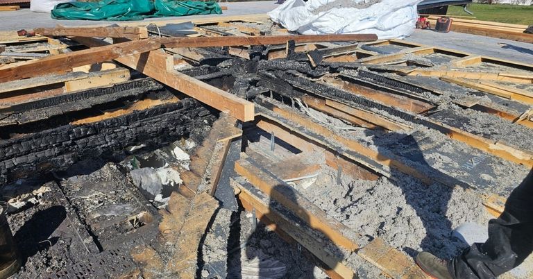 Das Bild zeigt eine Baustelle mit Holzbalken, einige verbrannt und andere intakt. Schutt und Staub bedecken das Gebiet. Oben links ist eine grüne Plane zu sehen.