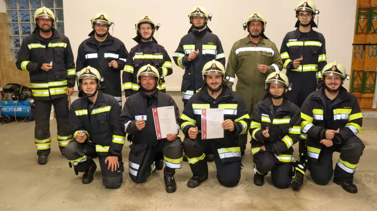 Eine Gruppe von Feuerwehrleuten in Uniform, einige knien und halten Urkunden in der Hand, posieren für ein Foto. Sie tragen alle Helme und Jacken mit gelben und schwarzen Streifen.