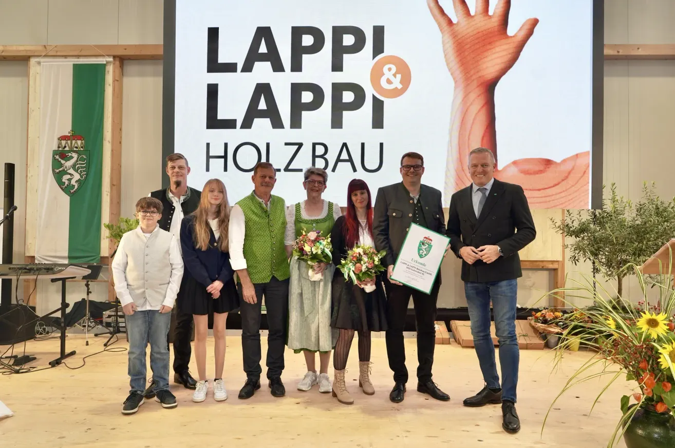 Eine Gruppe von Menschen posiert mit Blumen und einem Zertifikat vor einem großen Schild mit der Aufschrift LAPPI & LAPPI HOLZBAUE.