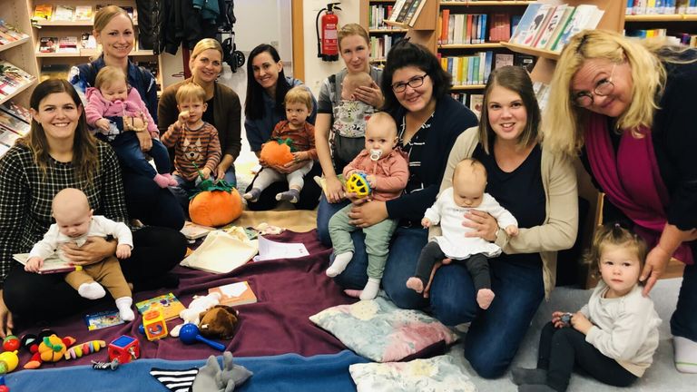 A group of women and children are sitting on a blanket in a library. Several babies are being held by their mothers, and there are pumpkins, books, and toys around them.