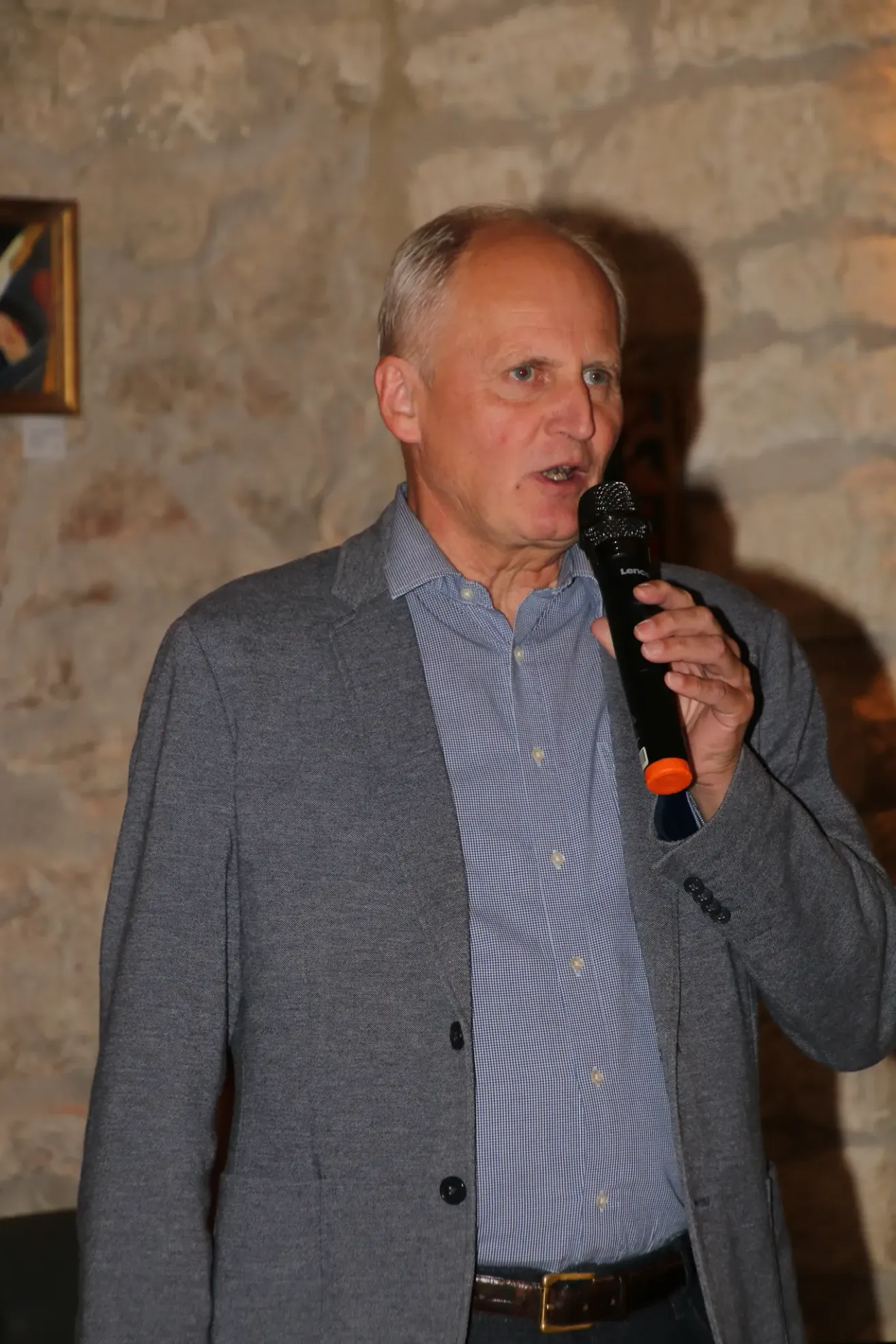Ein älterer Mann in einem grauen Blazer spricht in ein Mikrofon und steht vor einer Steinmauer.