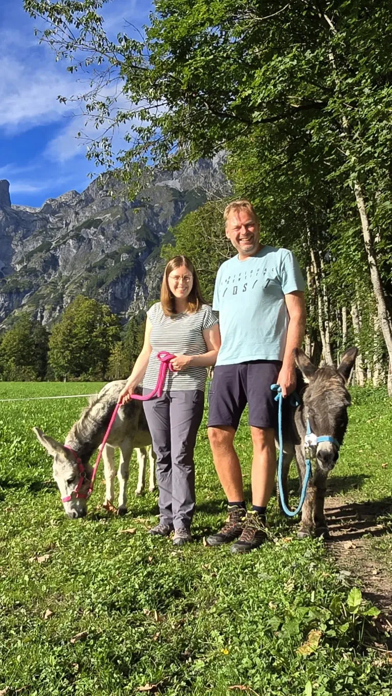 Ein Mann und eine Frau stehen auf einer Wiese mit zwei Eseln. Die Frau hält eine rosa Leine und der Mann eine blaue. Sie lächeln, mit Bergen im Hintergrund.