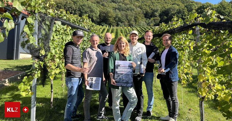 Sieben Menschen stehen in einem Weinberg mit Weingläsern und Flaschen in der Hand und lächeln für ein Foto. Eine Frau hält ein Plakat mit dem Titel 'Ortswein Prasentation'.
