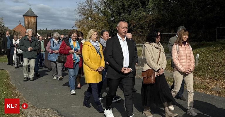 Eine Gruppe von Menschen, die draußen geht, einige in formeller Kleidung, andere in lässiger Kleidung. Sie gehen auf einem Weg mit einem Zaun an der Seite und Bäumen im Hintergrund.