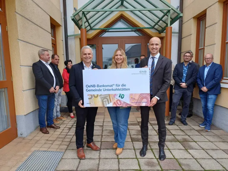 Drei Personen stehen vor einem Gebäude und halten ein großes Schild. Sie lächeln und schauen in die Kamera. Dahinter stehen weitere Personen, darunter ein Mann im Anzug.