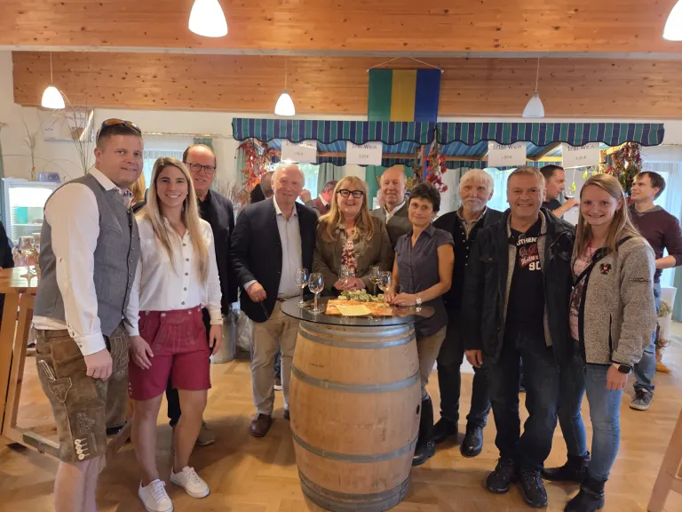 Eine Gruppe von Menschen versammelt sich um einen Holzfass, lächelt für ein Foto. Sie halten Weingläser und einige tragen Brillen. Ein Menü liegt auf dem Fass, und Flaggen hängen von der Decke.