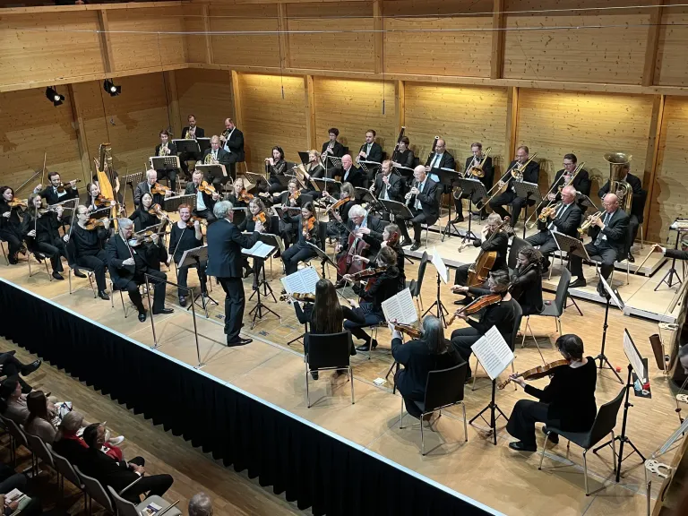 Ein Symphonieorchester tritt auf einer Bühne mit Holzboden auf. Mehrere Musiker sitzen, andere stehen und spielen ihre Instrumente. Das Publikum sitzt vorne.