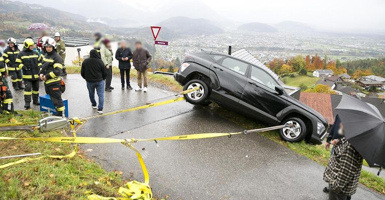 Ein schwarzer SUV wird auf einer nassen Straße abgeschleppt, vier Personen stehen in der Nähe. Das Fahrzeug ist mit gelben Seilen gesichert, im Hintergrund ist ein dreieckiges Schild sichtbar.