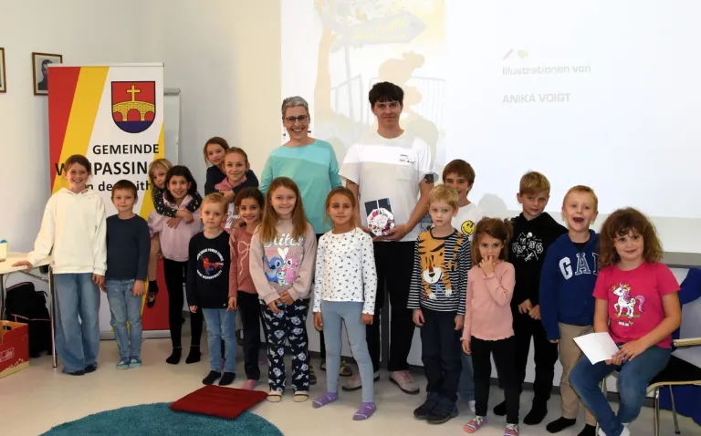 A group of children and two adults pose for a photo in a classroom. The children wear various clothing, and one holds a book. Behind them is a whiteboard with a logo and text.