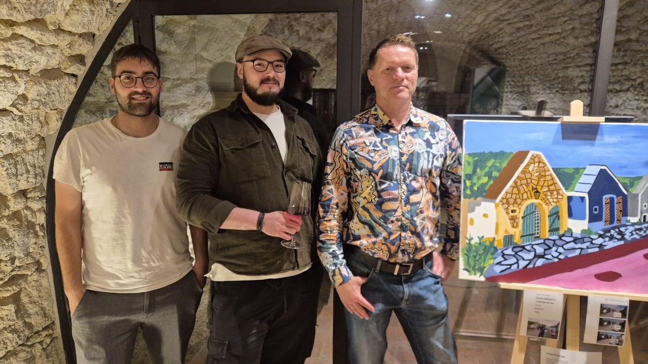 Drei Männer stehen eng beieinander vor einer Wand, einer mit einem Weinglas, ein anderer mit einem Hut und der dritte mit einem gemusterten Hemd. Hinter ihnen befindet sich ein Gemälde an der Wand.