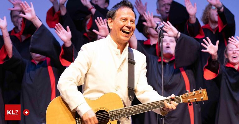 A man playing guitar and smiling while performing on stage with a choir behind him.