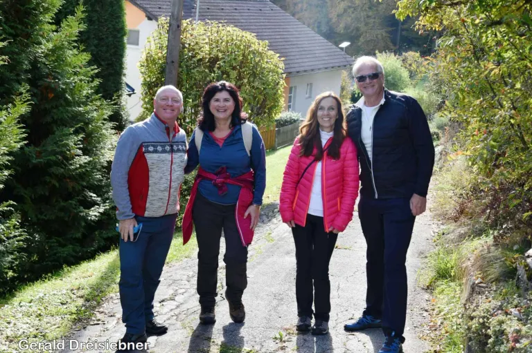 Vier Menschen stehen auf einem Pfad und lächeln für ein Foto. Sie sind alle für Outdoor-Aktivitäten gekleidet, wobei die Frau in Pink eine Jacke trägt. Sie stehen vor einem Haus mit Garten, mit Bäumen und Pflanzen im Hintergrund.