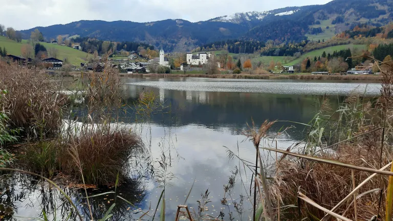 Ein ruhiger See spiegelt den Herbsthimmel und die nahen Berge wider. Dorfhäuser säumen die Landschaft, mit einem markanten weißen Kirchturm. Üppige Bäume und Sträucher säumen den Wasserrand.