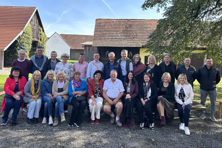 Eine Gruppe von lässig gekleideten Menschen, die auf einem Kiesweg vor einem Haus sitzen und stehen. Einige tragen Brillen und Schals. Das Haus hat eine gelbe Fassade mit einem braunen Dach. Bäume und Pflanzen sind im Hintergrund.