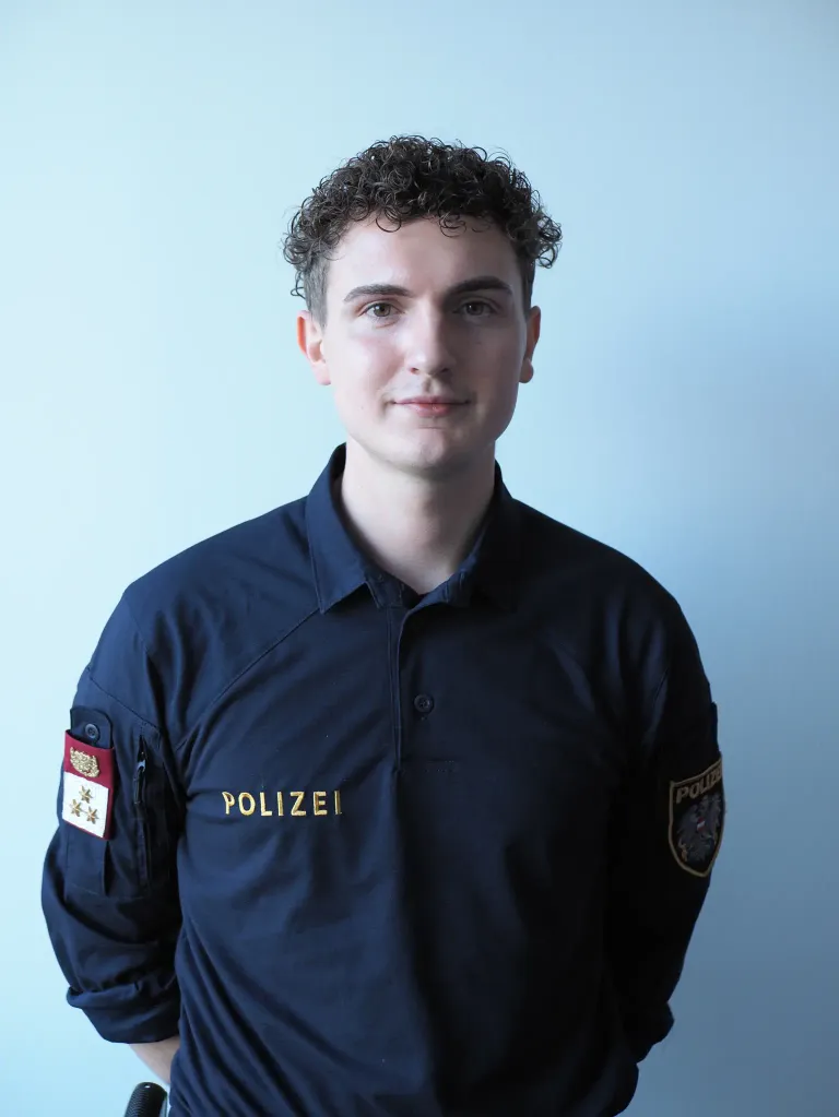 Ein Mann in einer blauen Polizeiuniform lächelt und posiert für ein Foto. Er hat lockige Haare und Abzeichen an seinen Ärmeln. Das Wort Polizei steht auf seinem Hemd.