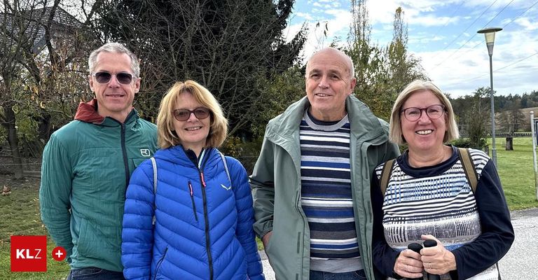 Vier Menschen stehen lächelnd eng beieinander vor einem Hintergrund aus Bäumen und blauem Himmel. Sie tragen Freizeitkleidung, was auf ein freundliches Treffen hindeutet.
