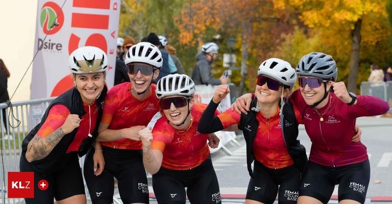 Fünf Frauen in Radbekleidung, mit Helmen und Sonnenbrillen, feiern auf der Straße, mit Herbstbäumen im Hintergrund.