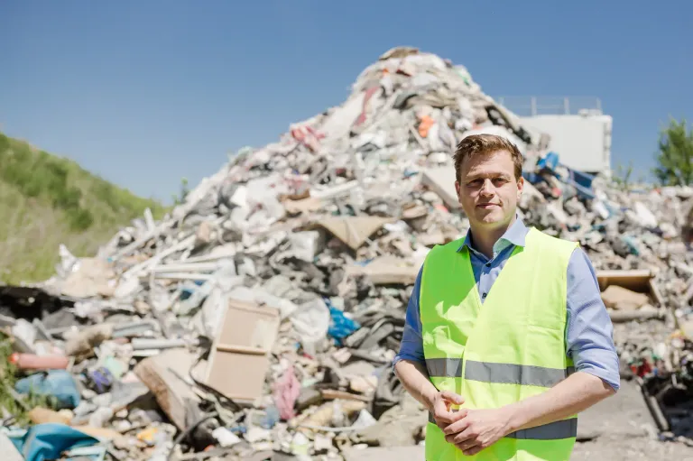 Ein Mann in einer Sicherheitsweste steht vor einem großen Haufen gemischter Abfälle. Er lächelt und scheint für ein Foto zu posieren.