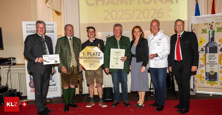 Eine Gruppe von Menschen steht auf einem roten Teppich, lächelt und posiert für ein Foto. Die beiden in der Mitte halten Auszeichnungen und Urkunden. Dahinter zeigt ein Banner 'CHAMPIONAT 2025/2026'.