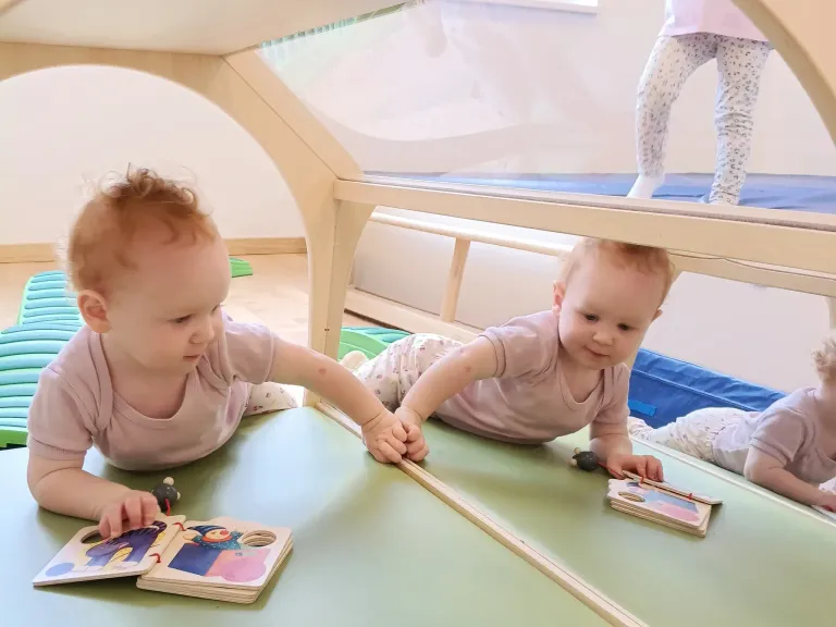 Zwei Säuglinge liegen auf einem Tisch, jeder hält ein Buch. Ein Baby hat einen Spielzeugwagen auf dem Tisch. Dahinter steht eine Person auf einer blauen Matte.