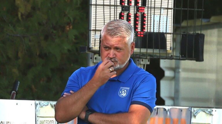 Ein Mann in einem blauen Shirt mit einem Logo auf der linken Brust, mit verschränkten Armen, einem Ring tragend und nach links schauend. Dahinter eine Metallbarriere mit roten Lichtern.