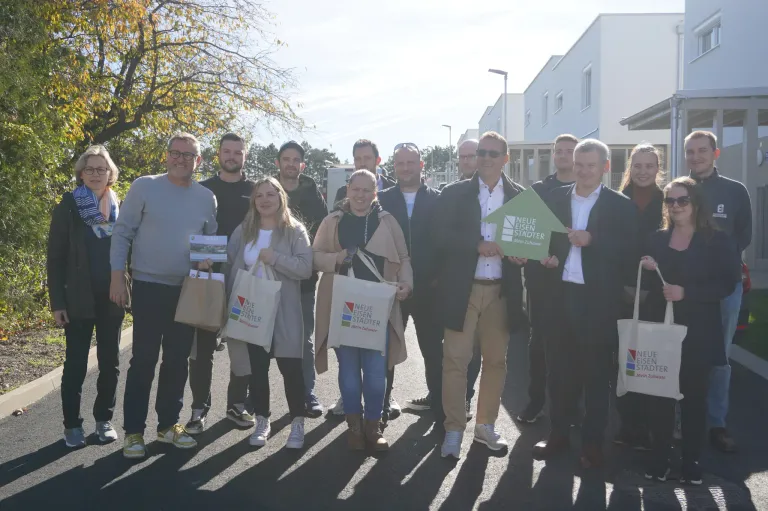 Eine Gruppe von Menschen posiert für ein Foto vor einem Gebäude. Sie alle halten Taschen und einige haben Bücher. Der Mann auf der rechten Seite hält ein grünes Schild.