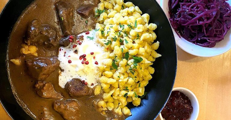 Eine Schüssel Rindergulasch mit cremigem Pilzrahm, überzogen mit roten Pfefferkörnern, serviert mit goldenen Kartoffelknödel, garniert mit Petersilie.
