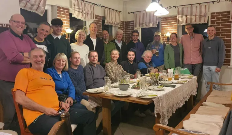 Eine Gruppe von Menschen versammelt sich um einen Tisch im Innenbereich, lächelt und posiert für ein Foto. Sie sind lässig gekleidet und scheinen eine Mahlzeit zu genießen. Der Tisch ist mit Tellern, Gläsern und Besteck gedeckt. Im Hintergrund befinden sich Ziegelwände und Lampen.